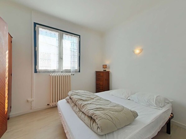 Un dormitorio con cama, un radiador, una ventana con cortinas, una lámpara de noche y un armario de madera.
