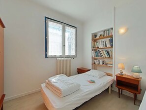 A small, neatly arranged bedroom with a single bed, a bedside table, a bookshelf, and a window with blinds.