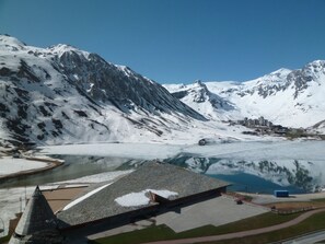 Paisagem montanhosa com um lago límpido, um edifício moderno com telhado inclinado e um pequeno povoado ao longe.