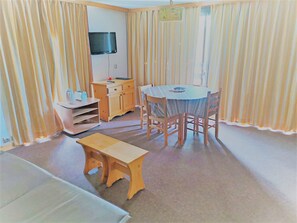 Una habitación con una mesa y sillas de madera, un mueble de madera pequeño, un televisor montado en la pared y una ventana con cortinas.
