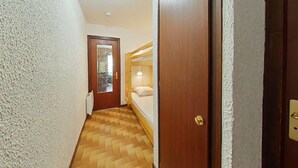 A narrow hallway with a wooden floor, a wooden door with a glass panel, and a bunk bed with a white mattress and a single pillow.