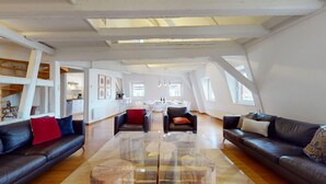 Ein großzügiges Loft mit hohen Decken, freiliegenden Balken und großen Fenstern. Es bietet einen Wohnbereich mit Ledersofas, einem Couchtisch und einen Essbereich mit Tisch für vier Personen.