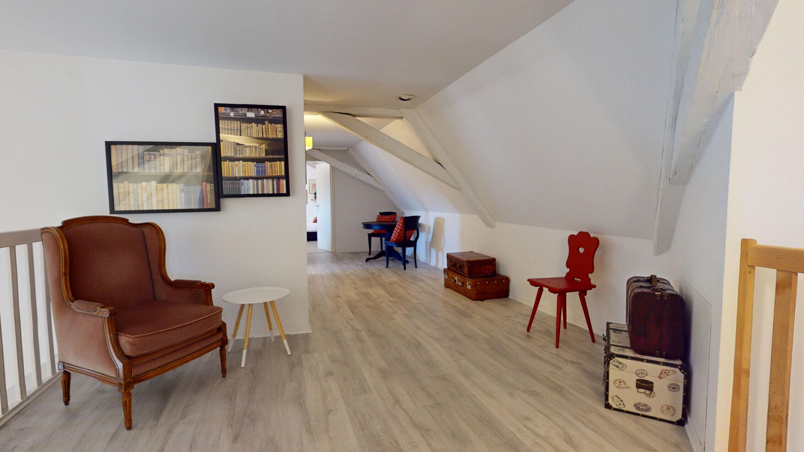 A loft-style living space with a high ceiling, exposed beams, and a sloped ceiling. There is a brown leather armchair, a small white table, and a red chair. A bookshelf and a suitcase are also visible.