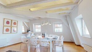Ein Essbereich mit weißem Tisch, gedeckt für sechs Personen, umgeben von weißen Stühlen, in einem Raum mit freiliegenden Holzbalken und großen Fenstern.