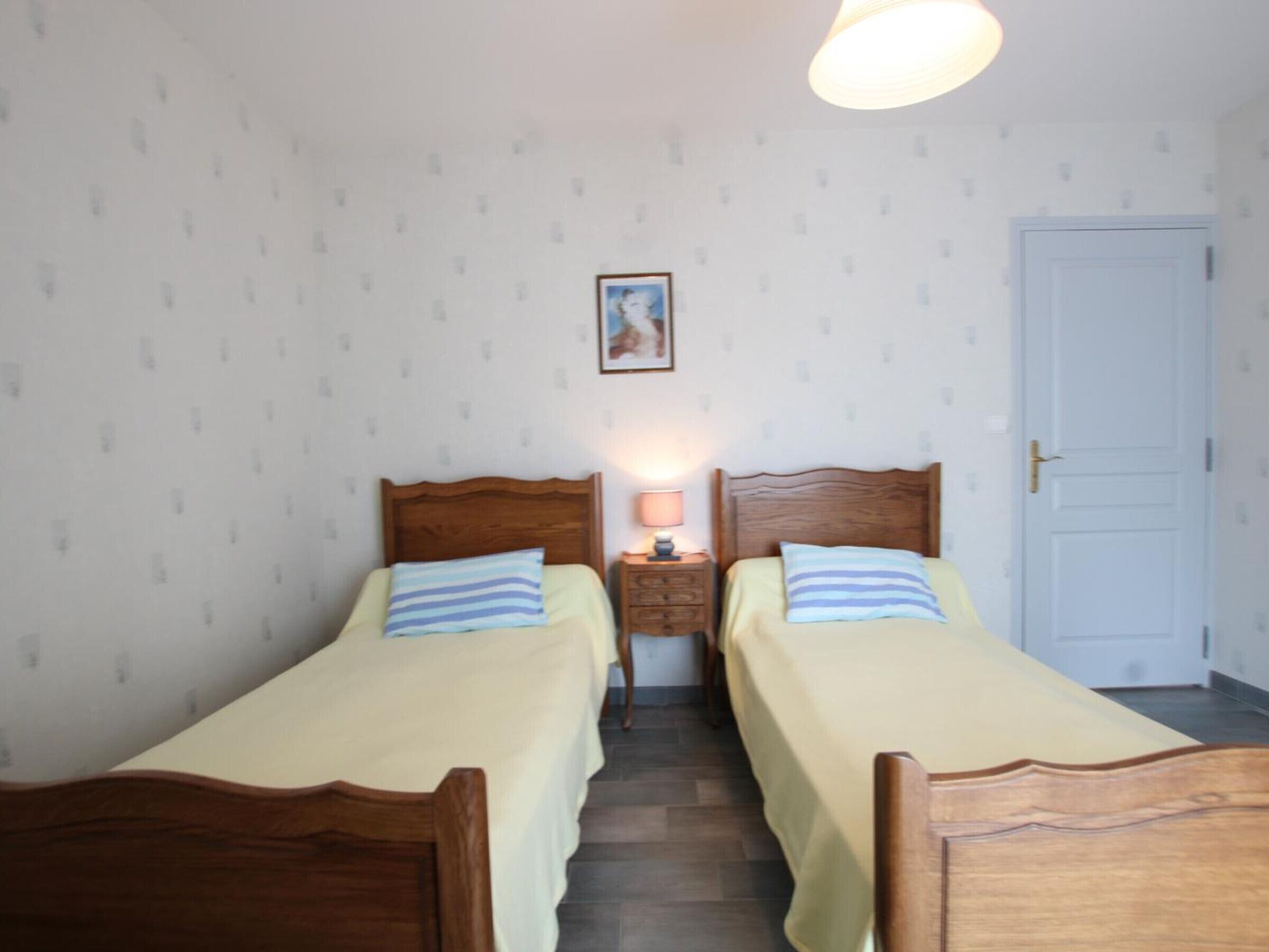 A room with two single beds, a nightstand with a lamp, a framed picture on the wall, and a white door.