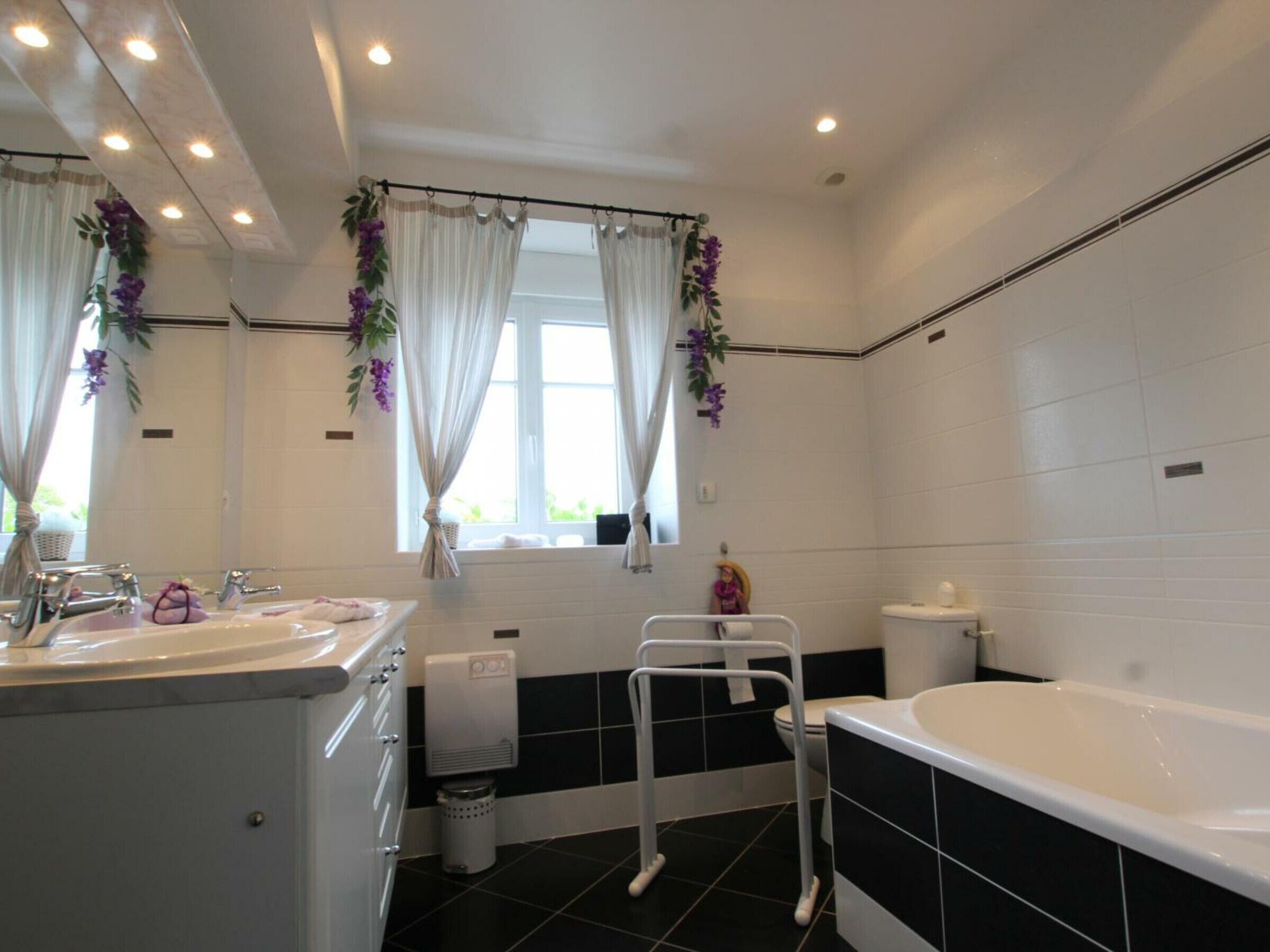 Un baño moderno con bañera, inodoro y lavamanos. Hay dos ventanas con cortinas y una pequeña planta en el alféizar.