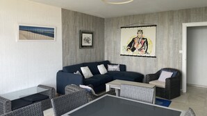 Ett modernt vardagsrum med en mörkblå soffa, fåtöljer i rotting, ett soffbord med glasskiva och konstverk på väggarna.