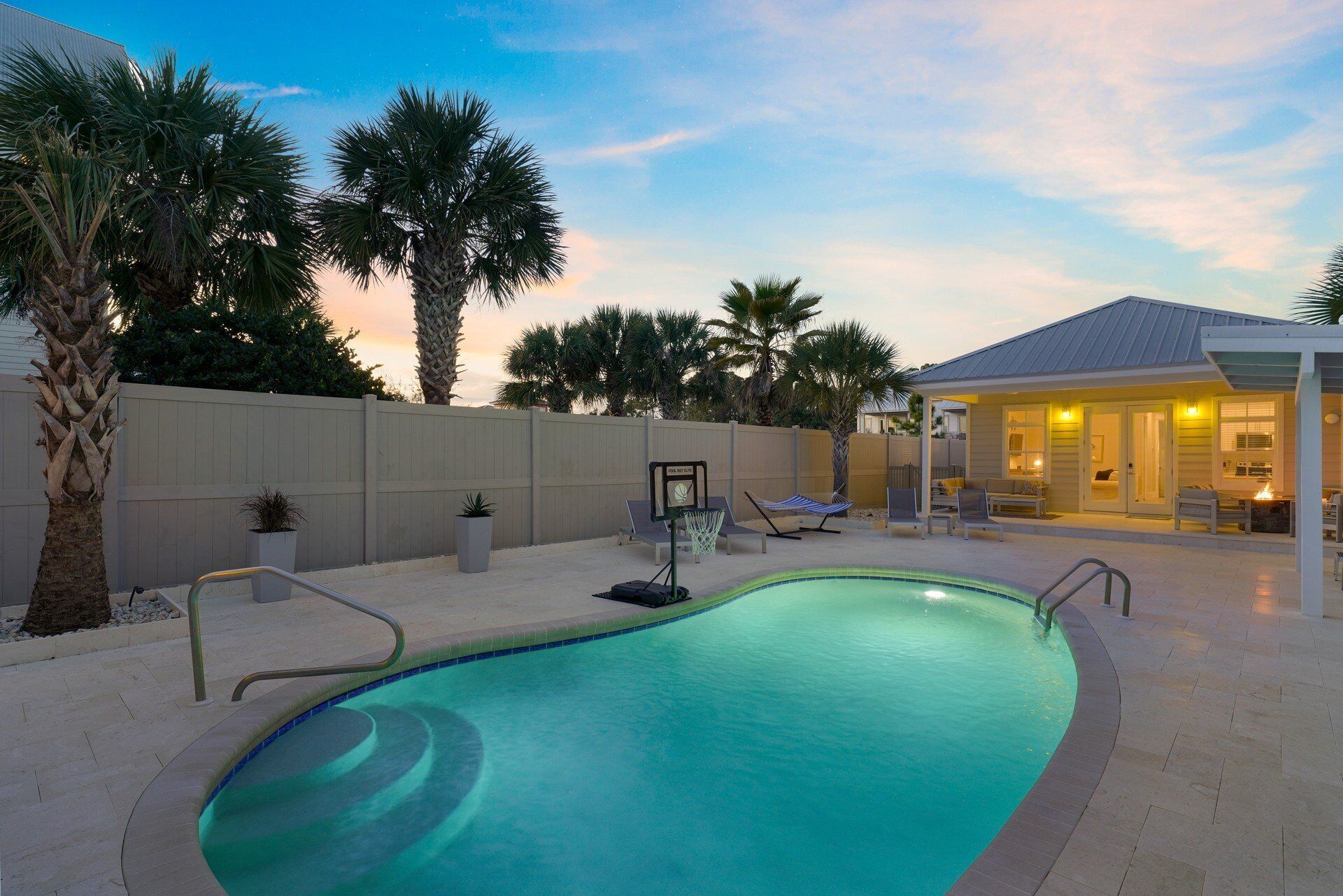 A swimming pool with a metal ladder, surrounded by palm trees and a covered patio area with seating.