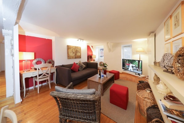 A living room with a sofa, coffee table, and a TV. There is a wicker chair and a red ottoman. The room has wooden flooring and white walls.