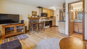 A modern kitchen with wooden cabinets, a dining area with a TV and speakers, and a cozy living room with a fireplace.