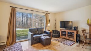 A living room with a leather sofa, a television, a cabinet with electronic devices, and a view of trees through large windows.