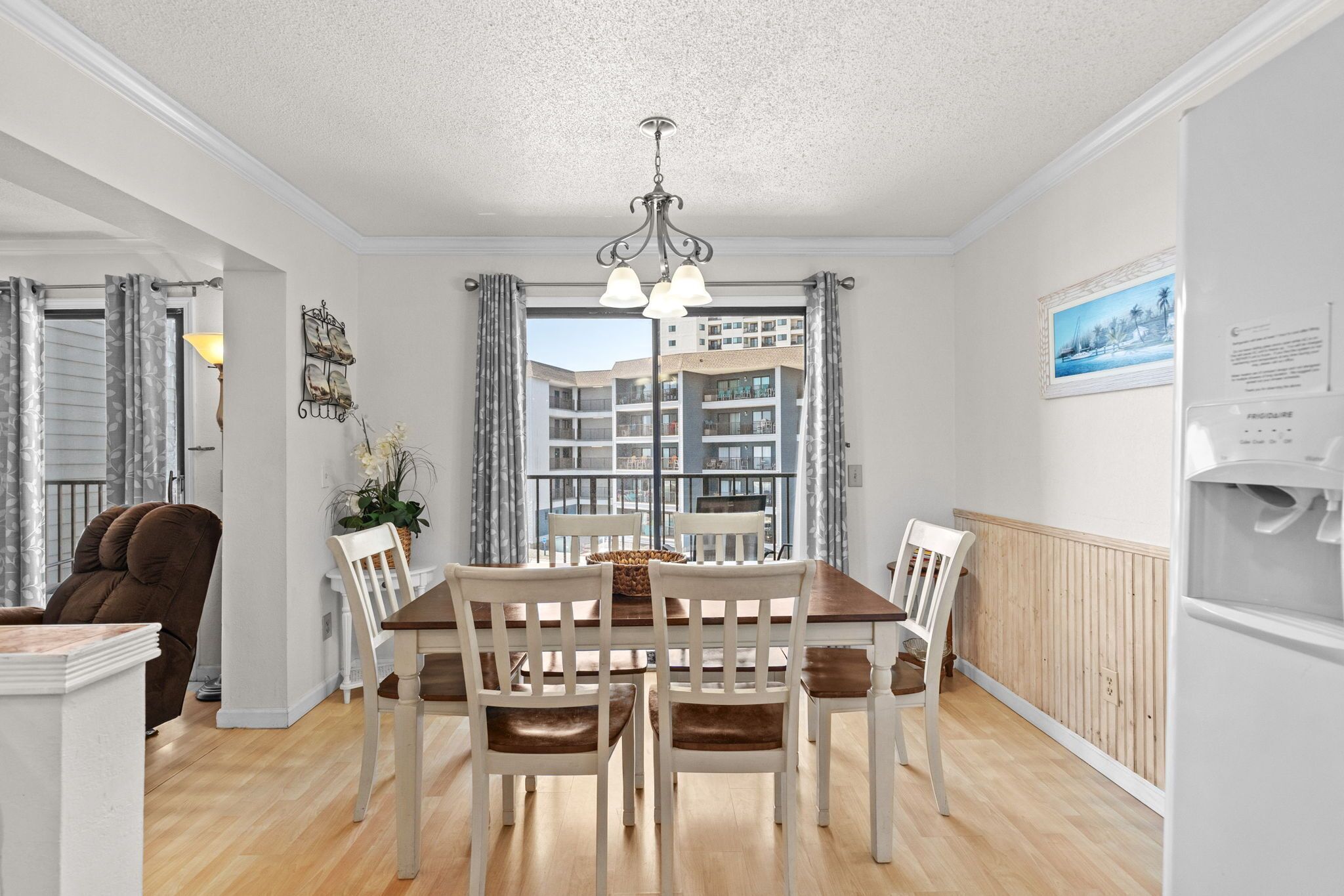 Comedor con mesa y sillas de madera, una araña y vistas a un edificio a través de amplios ventanales.