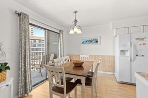 Comedor con mesa de madera, sillas blancas, lámpara colgante, refrigerador y vistas al balcón exterior.
