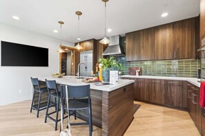A modern kitchen with wooden cabinets, a central island, bar stools, pendant lighting, and a large flat-screen TV.