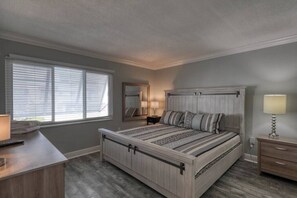 A bedroom with a large bed, a desk, a dresser, a mirror, and a window with blinds. - Coastal Condo · Renovated · Walk to Beach (U4) (Clearwater)