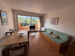 Quarto com mesa de jantar de madeira, sofá, televisão e varanda com vista para árvores e montanhas.