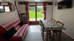 A cozy room with a sofa, a dining table with chairs, a TV, and a view of a green landscape through a door. - Cozy apartment with terrace for 4 people and parking in Châtel (Châtel)