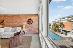 A hotel room with a brick wall, a bed, a wicker chair, a small table with a lamp, and a view of a pool and buildings outside. - This fabulous upscale condo is located right on the ship channel and in the hear (Port Aransas)