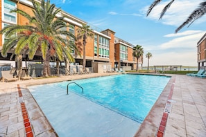 Una piscina con sillones de descanso y palmeras, rodeada por un edificio con varios pisos y balcones.