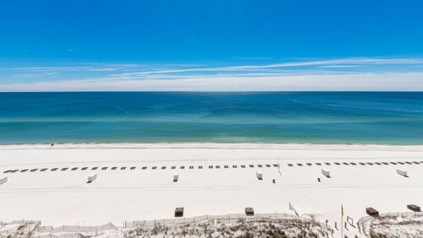 En strand med hvidt sand og en klar blå himmel. Der er rækker af strandstole og parasoller.