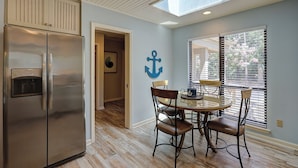 Una cocina con un refrigerador de acero inoxidable, un comedor con una mesa redonda y cuatro sillas, un adorno ancla en la pared y un tragaluz sobre el comedor.