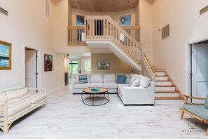 A spacious living area with a white sofa, a round coffee table, and a staircase with wooden railings. - 14 Bittern St - 5 Bedroom Home close to Ocean and Coligny (Hilton Head Island)