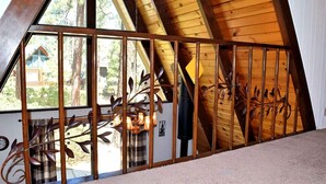 Una stanza con soffitto a falde, travi in legno e vista su una baita attraverso una finestra con ringhiera in metallo.