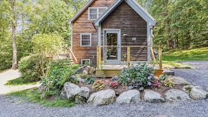 Une cabane en bois dotée d’un porche à l’avant, entourée d’un jardin agrémenté de rochers et de plantes.