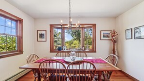 Une salle à manger avec une table en bois, des chaises, un lustre et de grandes fenêtres donnant sur un paysage verdoyant.