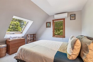 A bedroom with a skylight, a bed with multiple pillows, a wooden nightstand, a small table, and a window with a view of trees. - Ayers Farm Cottage: 4 br, wood fireplace, a/c, deck, mountain view! (Stowe)