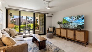 A modern living room with a sofa, coffee table, and TV, featuring a large window with a view of a balcony and outdoor furniture.