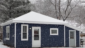 Una casa pequeña de un solo piso con fachada azul oscuro y molduras blancas. Tiene techo a dos aguas y una puerta principal con un pequeño ventanal encima. Está nevando, cubriendo el suelo y los árboles.