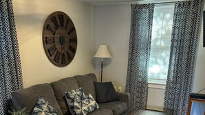 Una sala de estar con un sofá, una mesa de centro, un reloj de pared, una lámpara y cortinas estampadas.