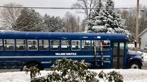 Un autobús azul con la leyenda "VILLAGE SHUTTLE" estacionado en una zona nevada, con árboles y una casa al fondo.