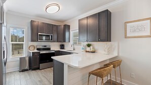 A modern kitchen with dark cabinets, a white countertop, a built-in microwave, and a stainless steel refrigerator. There are wicker barstools and a framed picture on the wall. - New Owners of Dock Knocker! Brand New Look In Time For Spring Break 2025! (Pensacola)