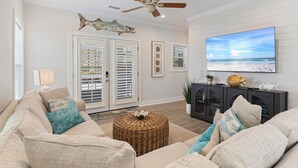 A living room with a large flat-screen TV, wicker ottoman, and a ceiling fan. - New Owners of Dock Knocker! Brand New Look In Time For Spring Break 2025! (Pensacola)