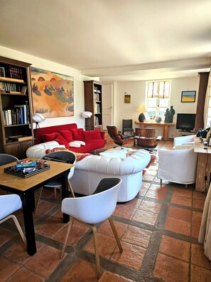 Un salon avec un canapé rouge, des fauteuils blancs, une table à manger et une bibliothèque.
