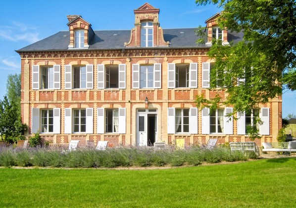 A large, two-story brick building with multiple windows and shutters, surrounded by a well-maintained lawn and trees.