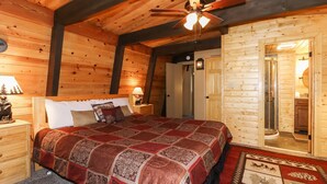 Ein Schlafzimmer in einer Holzhütte mit einem Bett, Nachttischen, einem Deckenventilator und einem durch eine offene Tür einsehbaren Badezimmer.