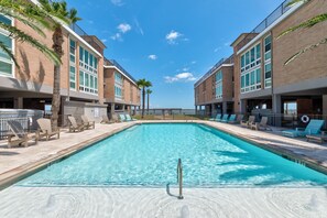 A rectangular swimming pool with a metal ladder, surrounded by lounge chairs and palm trees, adjacent to brick buildings with large windows. - Experience upscale coastal living in this fabulous condo with elevator access, p (Port Aransas)