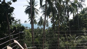 Una zona de sol con dos tumbonas y cojines, rodeada por una barandilla de metal, con vistas a un paisaje tropical con palmeras y vista al mar.