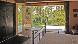 Un dormitorio con una cama grande, cabina de ducha de vidrio y balcón con vistas a palmeras.