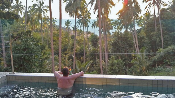 Un hombre en una piscina con vistas a altas palmeras y un cuerpo de agua al fondo.
