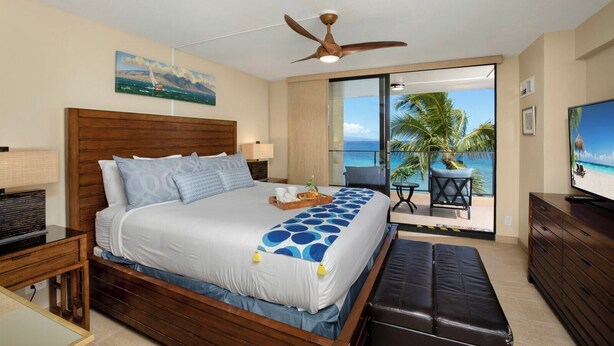 A hotel room with a bed, a TV, a ceiling fan, a balcony with a view of the ocean, and a painting on the wall. - Mahana Villas | Oceanfront 2 BR | Whale Watching | Car Incl. w/ 6+ Nights | MAH-611 by KBM (Lahaina)