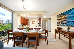 Un coin repas avec une table dressée pour quatre personnes, une cuisine en arrière-plan et une grande fenêtre donnant sur un paysage verdoyant et des bâtiments.
