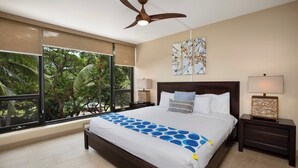 A bedroom with a large bed, a ceiling fan, a window with a view of trees, and artwork on the wall. - Mahana Villas | Oceanfront 2 BR | Whale Watching | Car Incl. w/ 6+ Nights | MAH-611 by KBM (Lahaina)