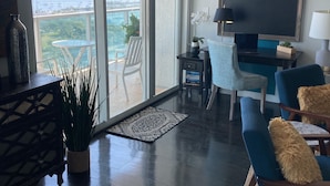 A modern living room with a large sliding glass door leading to a balcony with a view of the sea. There is a TV, a small table with a lamp, and two blue armchairs. - Luxury Penthouse w/Private Rooftop Terrace in Coconut Grove Hotel! (Miami)