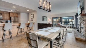 A modern kitchen with a bar area, a dining table with chairs, a stone fireplace, and a large window overlooking a balcony. - Luxury Ski-In Penthouse- Walk to Main St (Breckenridge)