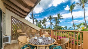 A round wooden table with four white mugs, surrounded by wicker chairs with blue cushions, on a balcony with a view of palm trees and a clear sky.