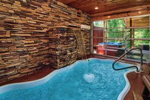 A hot tub with a waterfall feature, surrounded by a stone wall and wooden ceiling, with a view of outdoor seating through large windows.